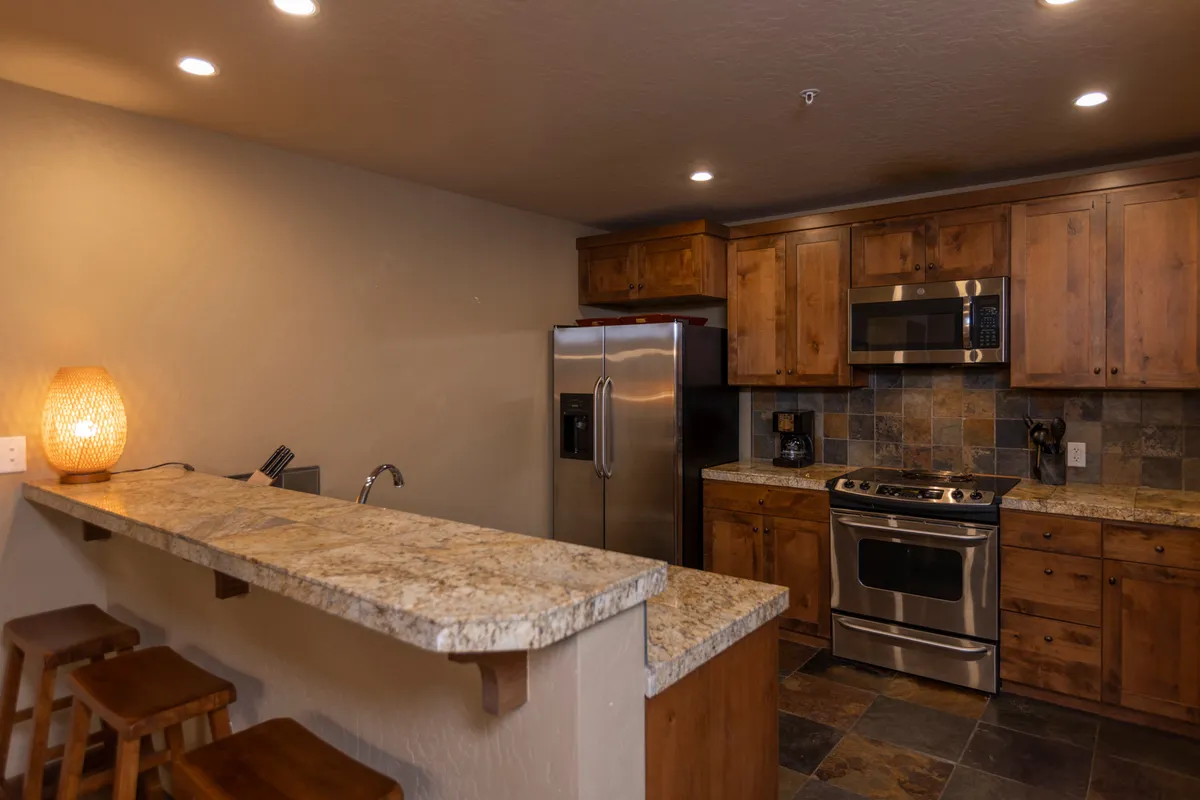This image captures the heart of the open-plan kitchen with a striking granite countertop, stainless steel appliances, warm wooden cabinetry, and bar stools. The ambient lighting suggests a cozy, inviting space perfect for cooking and socializing.