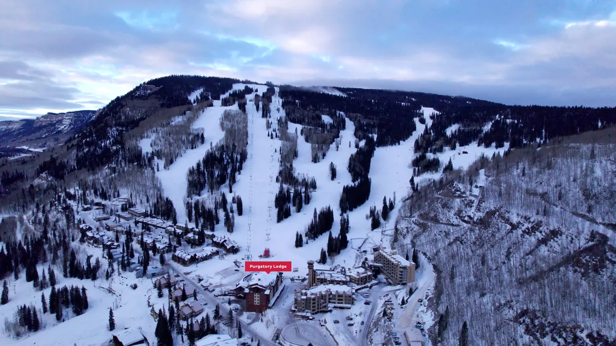 A sweeping exterior view of Purgatory Lodge nestled at the base of snow-covered ski slopes, with surrounding pine trees and mountains in the background. You can see the lodge's proximity to the slopes and the inviting atmosphere for skiers and snowboarders heading to and from the lifts.