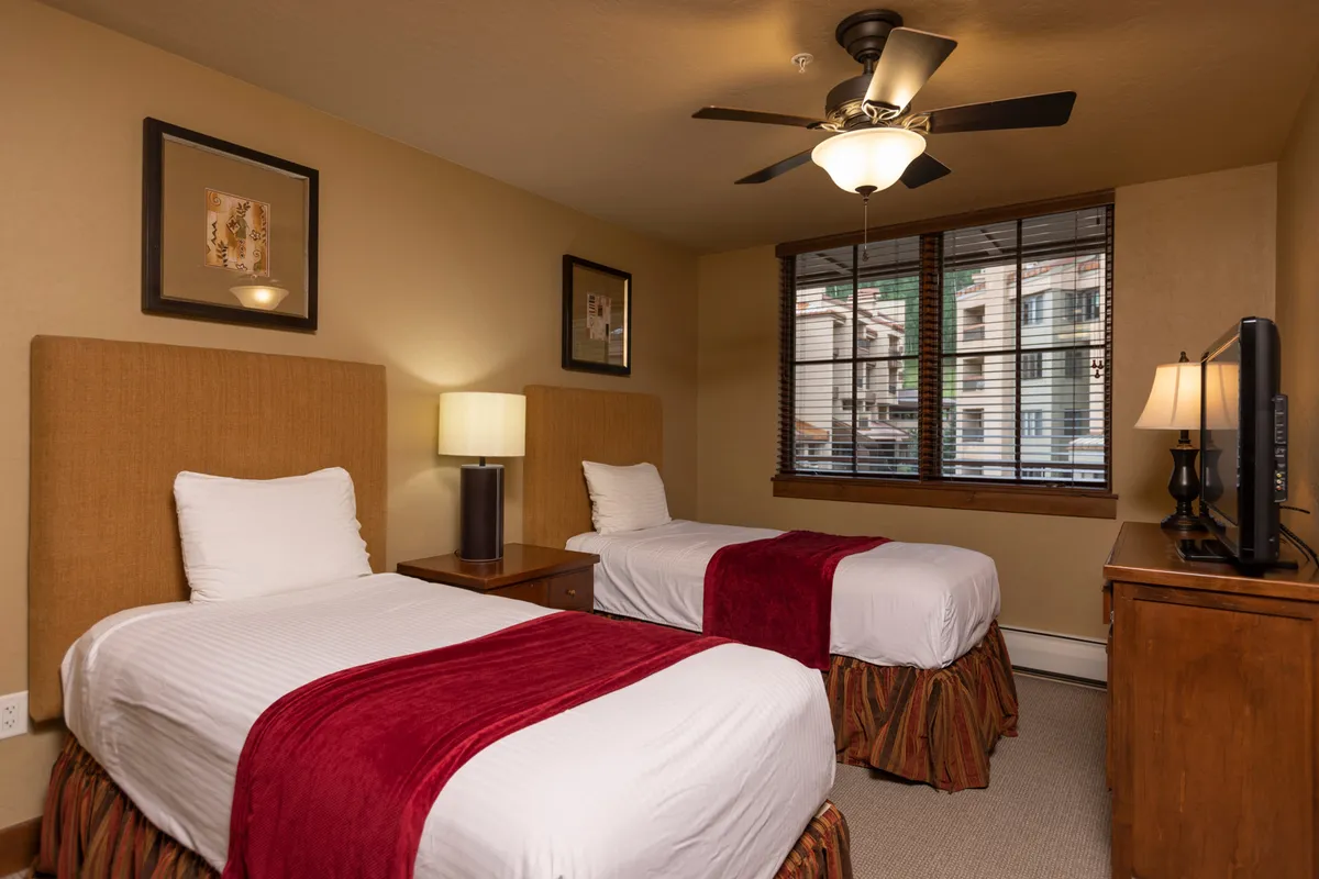 A bright bedroom with two twin beds adorned with red accent throws, a window with blinds letting in natural light, and a relaxed atmosphere ideal for families or friends sharing the space.