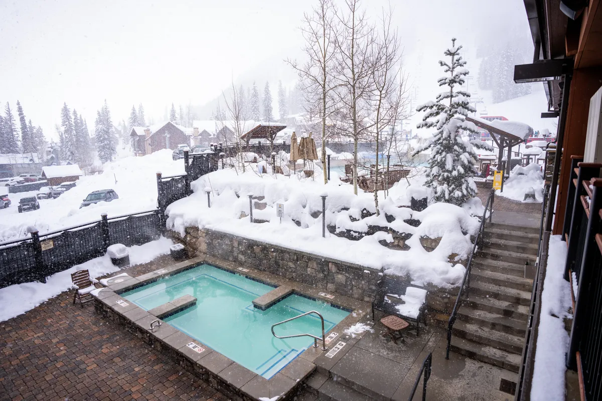 An outdoor hot tub nestled in a snowy landscape, surrounded by trees. The steam rises into the cool air as people soak, enjoying the serene winter scenery. Steps lead down to the tub, and a sense of tranquil luxury prevails.