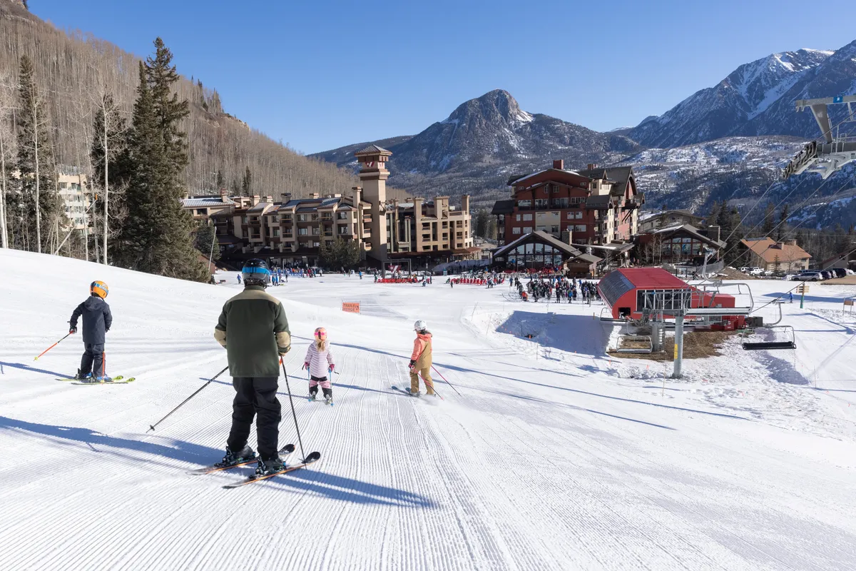 A vibrant exterior scene with skiers and snowboarders gliding down the slope, the mountain backdrop framing the lodge in the distance. Family-friendly energy is evident, with guests actively engaged in winter sports right outside the property.