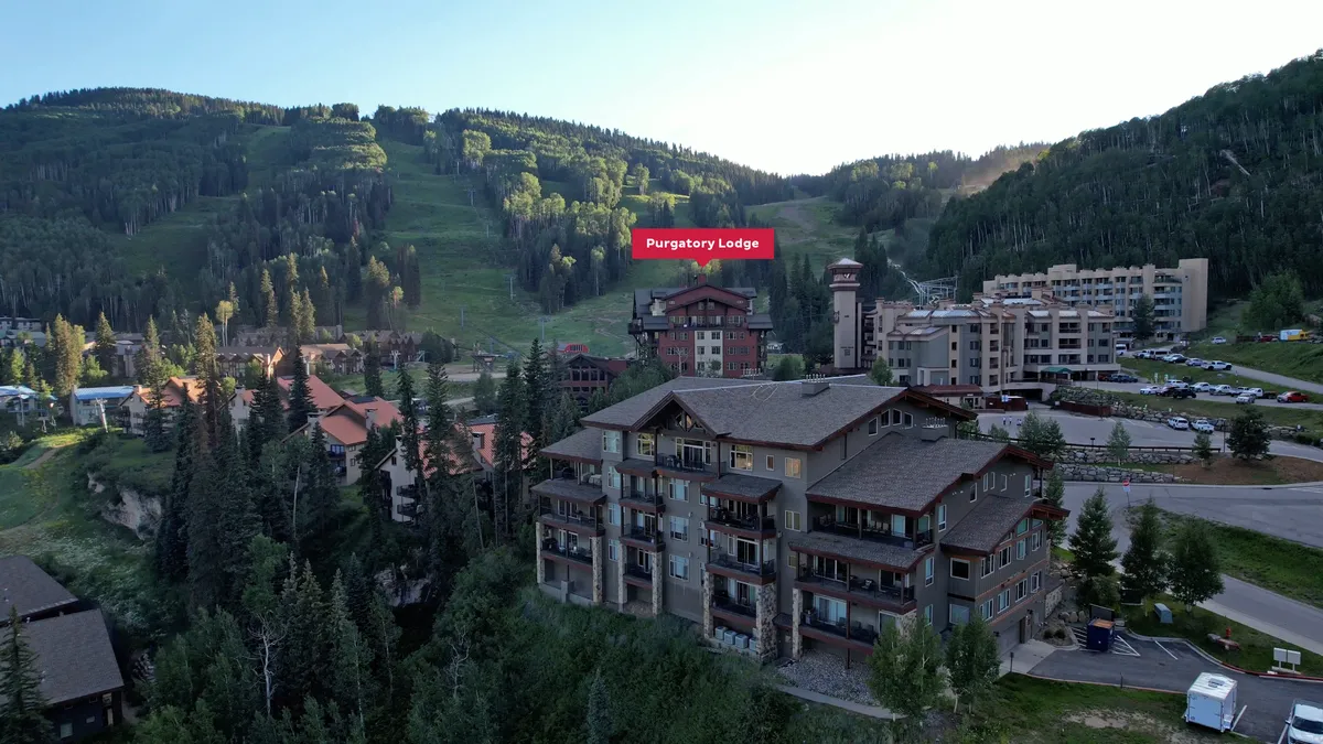 This exterior image features Purgatory Lodge set against a stunning mountain backdrop, surrounded by snow-capped peaks and adjacent buildings. The scene suggests immediate access to the slopes and captures the adventurous essence of ski-in/ski-out living, with the lodge as a cozy and welcoming base for mountain activities.