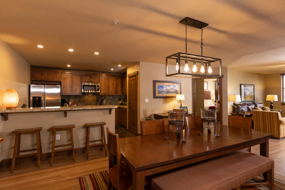The image showcases a spacious, warmly lit open-concept interior with a large dining table, a stylish kitchen island topped with granite, elegant lighting fixtures, and a plush sofa—perfect for gathering after a day on the slopes. The scene radiates coziness and invites guests to relax, dine, and enjoy quality time together in a luxurious setting.