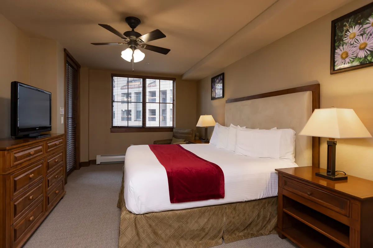 A cozy bedroom featuring a comfortable bed with a vibrant red throw, a wooden dresser, nightstand with a lamp, and a television. Morning light seeps in through a window, emphasizing the room's inviting ambiance.