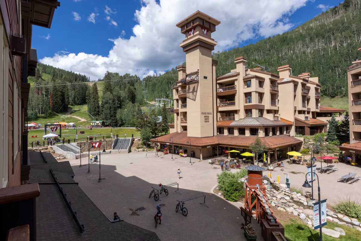This lively exterior scene features the building's architecture set against a dramatic mountain landscape, with guests enjoying the outdoor seating area, bicycles, and general leisure activities. The area buzzes with activity, hinting at the lively Purgatory Village and its events or gatherings just steps from the lodge.