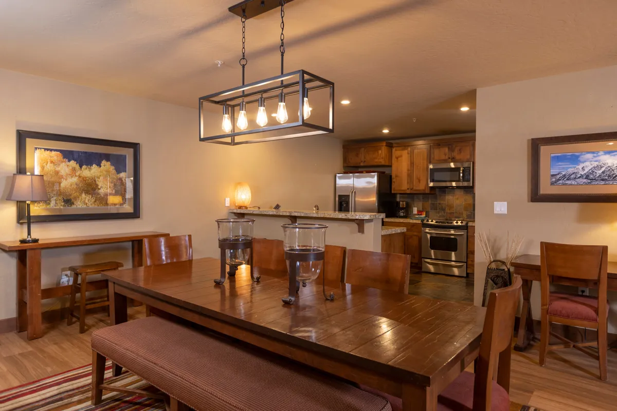 A warmly lit dining area featuring a stylish chandelier, a spacious dining table set for a meal, and an open view into the kitchen area. The setting is inviting, with touches like intricate artwork on the wall and soft candlelight creating a cozy, social atmosphere.