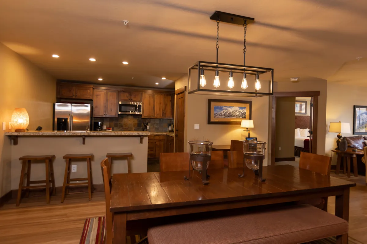 A bright, open-plan dining and kitchen area with a central dining table, a kitchen island with bar stools, and modern light fixtures. Artwork adds character to the space, and the overall mood is warm and welcoming, perfect for socializing and entertaining.