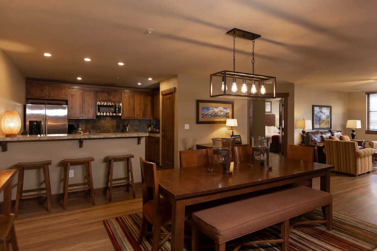 A wide view encompassing the dining table, adjacent bar stools, kitchen counter, and a comfortable sofa area, all bathed in warm, welcoming light. Shared spaces blend seamlessly, inviting gatherings for meals, relaxation, and conversation.